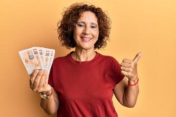 Poster - Beautiful middle age mature woman holding 10 united kingdom pounds banknotes smiling happy and positive, thumb up doing excellent and approval sign