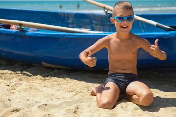 Wall Mural - A young boy in a blue boat on the ocean. A child in sunglasses on the beach near the shore.