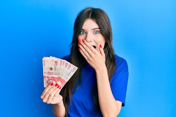 Sticker - Young beautiful caucasian girl holding 10 colombian pesos banknotes covering mouth with hand, shocked and afraid for mistake. surprised expression