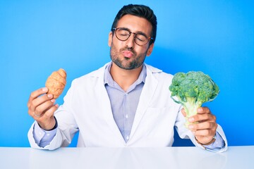 Young hispanic man as nutritionist doctor holding croissant and broccoli looking at the camera blowing a kiss being lovely and sexy. love expression.