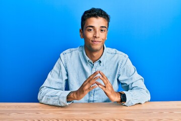 Young handsome african american man wearing casual clothes sitting on the table hands together and fingers crossed smiling relaxed and cheerful. success and optimistic