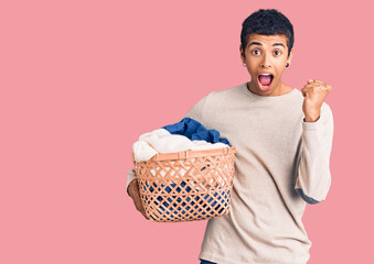 Wall Mural - Young african amercian man holding laundry basket screaming proud, celebrating victory and success very excited with raised arms