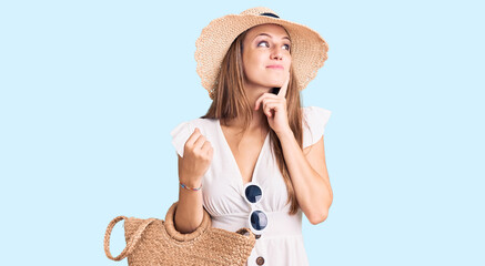 Wall Mural - Young beautiful blonde woman wearing summer dress and wicker handbag serious face thinking about question with hand on chin, thoughtful about confusing idea