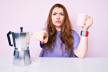 Poster - Young redhead woman sitting on the table drinking coffee pointing with finger to the camera and to you, confident gesture looking serious