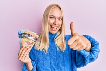 Wall Mural - Young blonde girl holding canadian dollars approving doing positive gesture with hand, thumbs up smiling and happy for success. winner gesture.