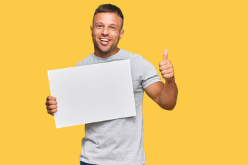 Poster - Handsome muscle man holding blank empty banner smiling happy and positive, thumb up doing excellent and approval sign