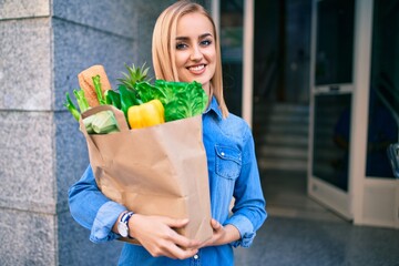 Young blonde girl smiling happy holding groceries paper bag standing at the city.