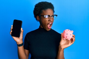 Sticker - Young african american girl holding piggy bank and smartphone angry and mad screaming frustrated and furious, shouting with anger. rage and aggressive concept.