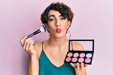 Young man wearing woman make up holding makeup brush and blush looking at the camera blowing a kiss being lovely and sexy. love expression.