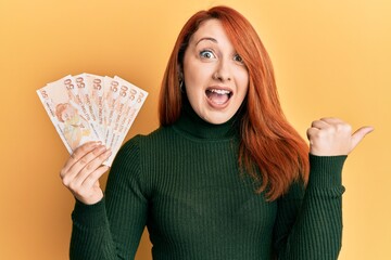 Wall Mural - Beautiful redhead woman holding 50 turkish lira banknotes pointing thumb up to the side smiling happy with open mouth