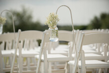 Fancy White Folding Chairs Lined  Up At Outdoor Wedding Ceremony With Babyie's Breathe Flowers In Mason Jars Hanging