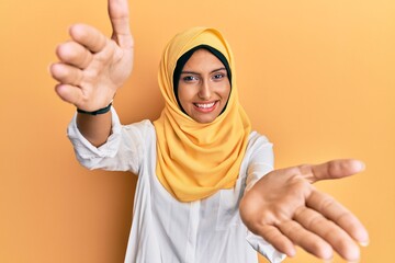 Poster - Young brunette arab woman wearing traditional islamic hijab scarf looking at the camera smiling with open arms for hug. cheerful expression embracing happiness.