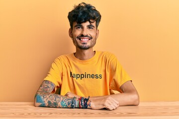 Wall Mural - Young hispanic man wearing tshirt with happiness word message sitting on the table with a happy and cool smile on face. lucky person.