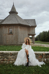 Canvas Print - Beautiful leggy model girl in a wedding dress and straw hat posing near the wooden church in the countryside. Young woman bride in a modish vintage dress. Concept of wedding fashion