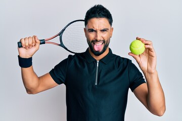 Young man with beard playing tennis holding racket and ball sticking tongue out happy with funny expression.