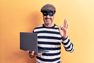 Middle age handsome burglar man wearing cap and mask using laptop over yellow background doing ok sign with fingers, smiling friendly gesturing excellent symbol