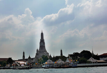 The high tower of Wat Arun against the blue sky. The pagoda is decorated with carvings, ornaments. In the foreground is the river, the pier. Bangkok. Thailand.