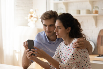 Wall Mural - Millennial Caucasian man and woman sit at home kitchen speak on video call or have webcam talk on smartphone. Young couple spouses use cellphone look at screen shopping on internet on gadget.