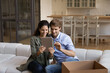 © fizkes - Happy millennial couple sit on sofa relax on moving day to new home speak talk on video call on tablet. Smiling young man and woman use pad gadget browsing internet in own house or apartment.