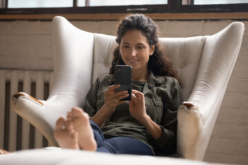 Poster - Smiling millennial Caucasian woman sit relax in cozy armchair in living room text or message online on cellphone. Happy young female rest in chair at home browse internet on cellphone gadget.