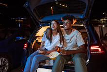 Diverse Young Couple Having Romantic Date. Cheerful Guy And His Girlfriend Watching A Movie, Sitting Together In Car Trunk In Front Of A Screen In An Open Air Cinema