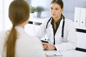 Wall Mural - Woman-doctor and patient sitting and talking at hospital office. Green color blouse suits to therapist. Medicine and healthcare concept