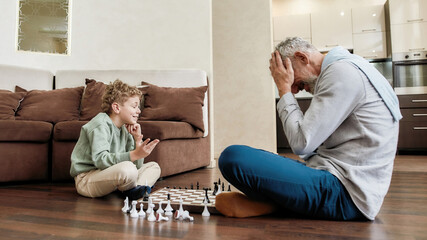 Grandfather and grandson playing chess together at home, sitting on the floor in the living room, middle aged man holding head in hands with confused face expression