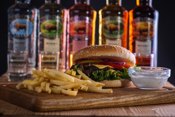 burger with baked slices of fried meat, tomatoes, onions, cheese, salad and french fries on a wooden surface, isolated on a dark background, in the background a bottle of alcohol