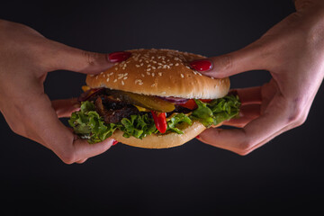 Girl holding a hamburger with two hands on a black background. baked slices of roasted meat, tomato, onion, cheese, salad. Copy space, on dark background. Delicious burger concept. There is an inscrip