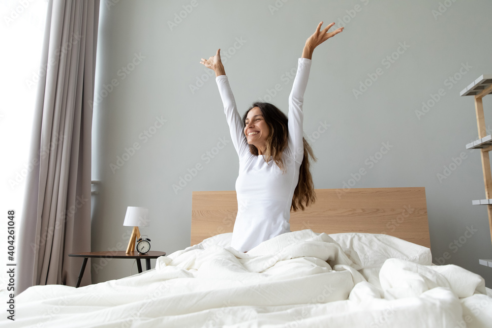 Overjoyed woman wearing pajama stretching hands after awakening, sitting in cozy bad, happy smiling attractive young female starting new day, enjoying morning after healthy sleep, doing exercises - obrazy, fototapety, plakaty 