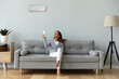 © fizkes - Smiling woman using air conditioner remote controller, sitting on cozy couch at home full length, happy young female relaxing setting comfort temperature in modern living room, enjoy fresh air