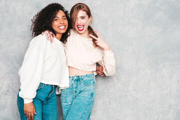 Two young beautiful smiling international hipster female in trendy summer jeans clothes. Sexy carefree women posing near gray wall in studio. Positive models having fun. Concept of friendship