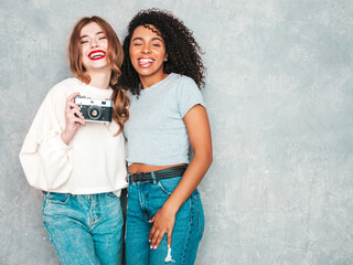 Two young beautiful smiling international hipster female in trendy summer jeans clothes. Sexy carefree women posing near gray wall in studio. Positive models having fun. Taking photos on retro camera