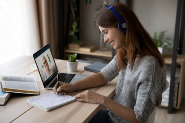 Smiling millennial female student in earphones make notes have online webcam lesson with teacher or tutor on computer. Happy young woman study on internet, talk on video call with coach on laptop.