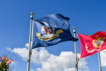 A Flag Of The United States Navy Flies Alongside And American Flag And A U.S. Marine Flag For The Armed Services In Front Of A Blue Sky.