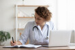 © fizkes - Concentrated young female general practitioner physician writing notes in registry book, working in clinic office. Skilled professional doctor prescribing disease treatment, involved in paperwork.