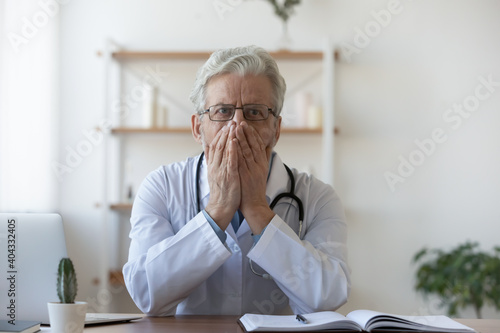 Confused old mature doctor therapist in glasses and white coat covering mouth with hands, thinking of medical mistake or wrong patient's diagnosis, feeling stressed and fearful in clinic office.
