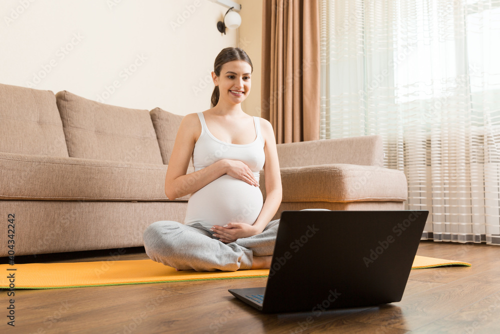 Young happy healthy yoga fitness beautiful pregnant woman doing bakasana yoga workout on yoga mat opposite laptop with online master class. lifestyle concept at coronavirus time - obrazy, fototapety, plakaty 