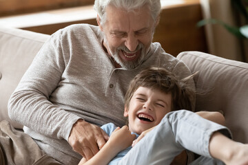 Overjoyed elderly grandfather have fun play tickle cute little 6s grandson, relax together on home weekend. Happy loving mature grandparent feel playful laugh and joke enjoy time with small boy child.