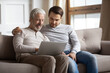 © fizkes - Happy millennial man embrace elderly father relax together in living room browsing internet on laptop. Smiling loving adult son rest enjoy family weekend time with mature dad using computer.