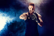 Bearded tattooed sportsman muay thai boxer in black undershirt and boxing gloves screams, motivates on dark background with smoke. Sport concept.