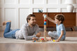 © fizkes - Caring young Caucasian father and small son sit on warm floor at home engaged in funny game together. Loving dad and little boy child have fun play build construct with wooden blocks bricks.
