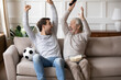 © fizkes - Excited millennial Caucasian man with elderly father feel euphoric celebrate win watch football game on TV together. Overjoyed adult son with mature dad triumph with team victory enjoy sports match.