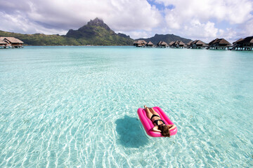 Woman floats on a pink inflatable mattress over beautiful clear, turquoise, tropical island ocean water while on vacation in Bora Bora, French Polynesia