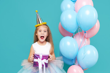 A joyful child girl in an elegant tulle dress and a festive cap holds a gift box and laughs, near the balloons. Birthday present. Funny face on cyan background