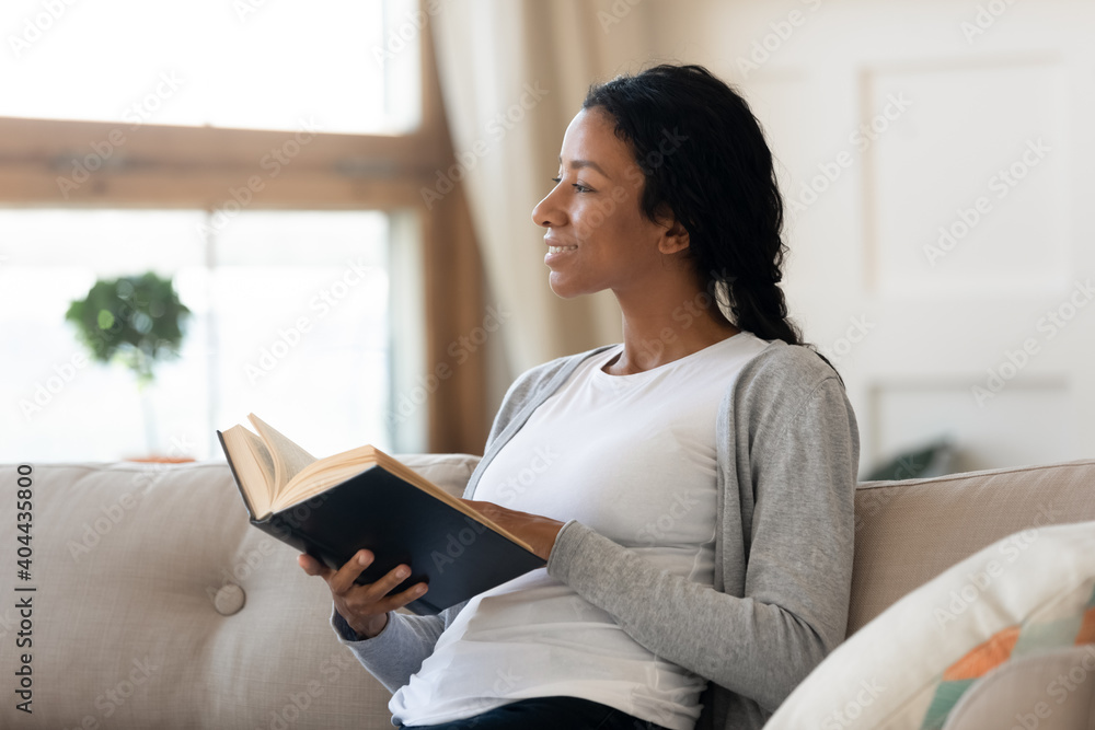 Dreamy young african american woman relaxing on comfortable sofa with paper book in hands, looking in distance. Happy millennial multiracial lady enjoying hobby time alone indoors, imagination concept - obrazy, fototapety, plakaty 
