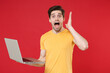 © ViDi Studio - Young shocked surprised unshaved successful caucasian student man 20s wearing casual basic yellow t-shirt using laptop pc computer spreading hands isolated on red color background studio portrait