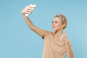 Young smiling blonde caucasian woman 20s short haircut dental braces in casual beige t-shirt doing selfie shot on mobile phone showing thumb up gesture isolated on blue background studio portrait