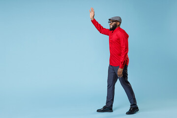 Wall Mural - Full length side view joyful young bearded african american man in casual red shirt cap eyeglasses waving and greeting with hand as notices someone isolated on pastel blue background studio portrait.