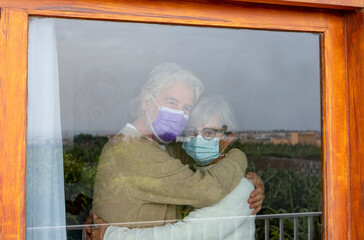 Two sad senior people stay at home behind the window for lockdown due to coronavirus. Elderly couple hug each other with love